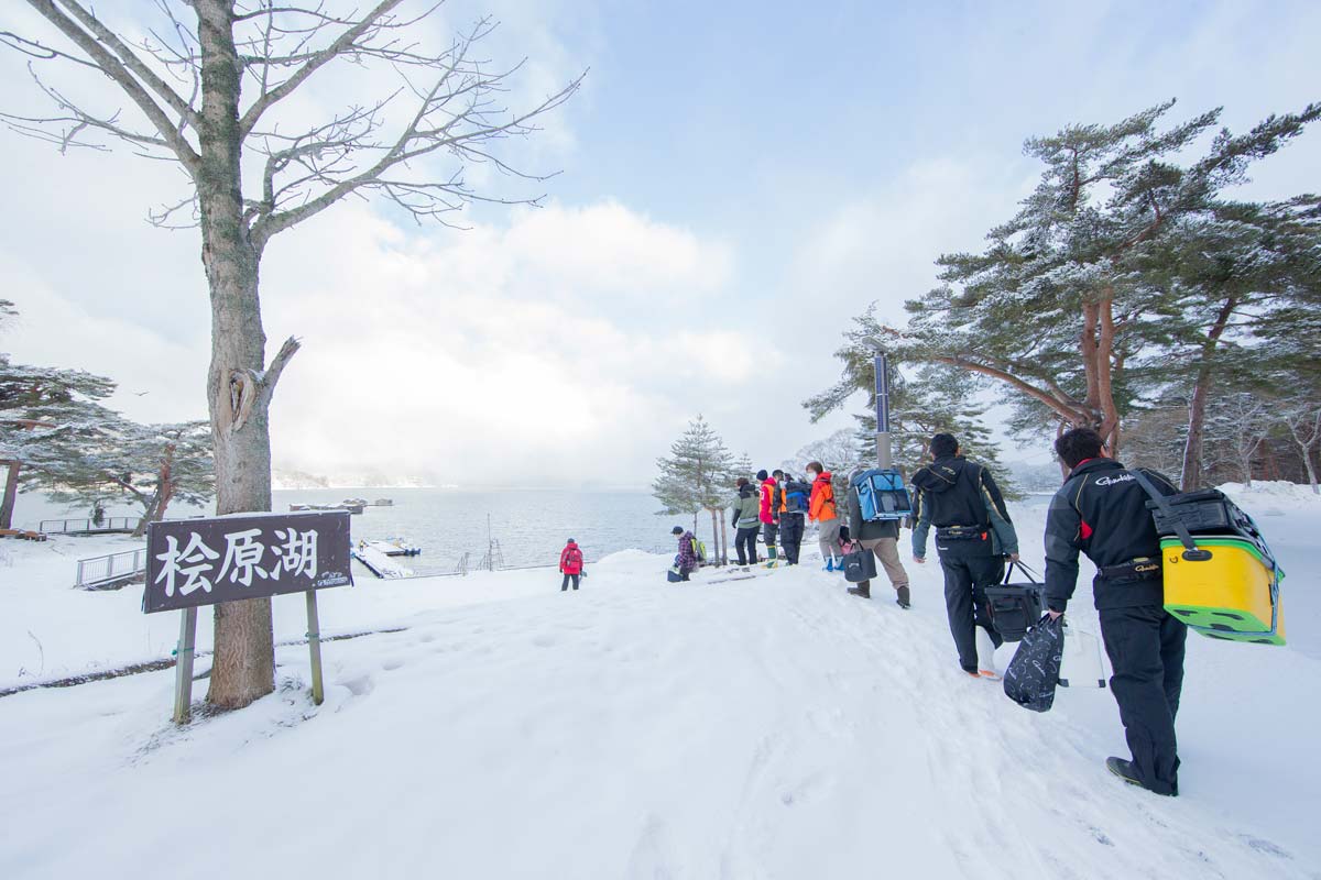 桧原湖の立て看板と、雪道を進み桧原湖へと向かう釣り人の列